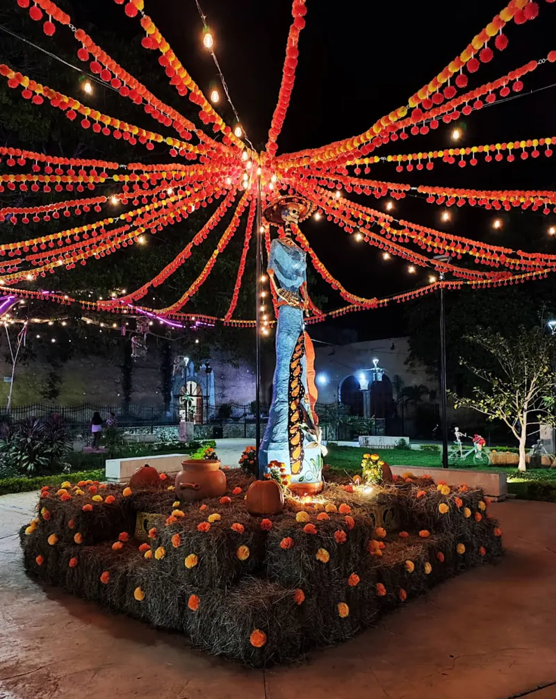 La plaza de noche bajo las luces de Hanal Pixán, con una catrina, flores de cempasúchil y la iglesia al fondo.